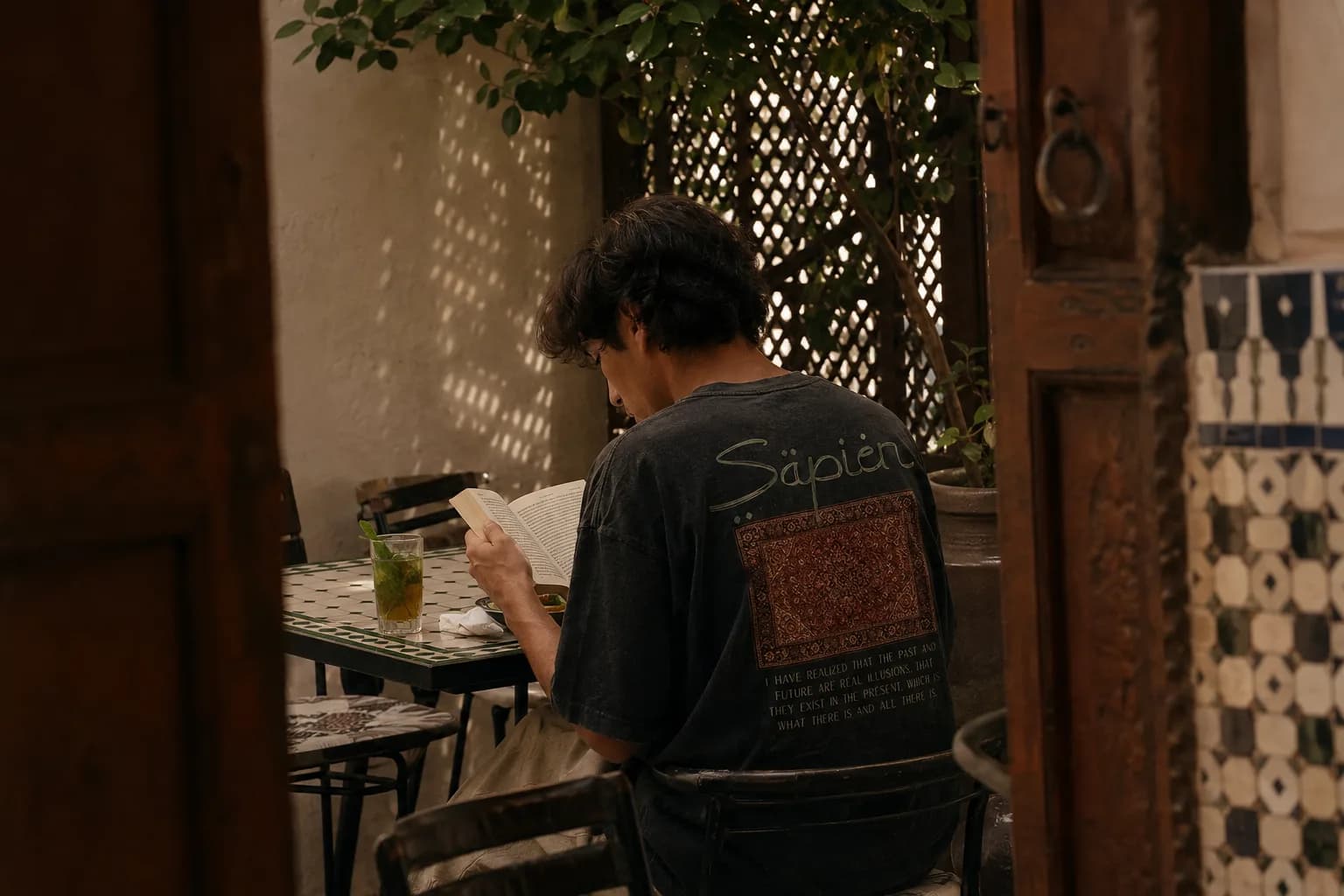 A man reads at a tiled courtyard table wearing the Alan Watts tee