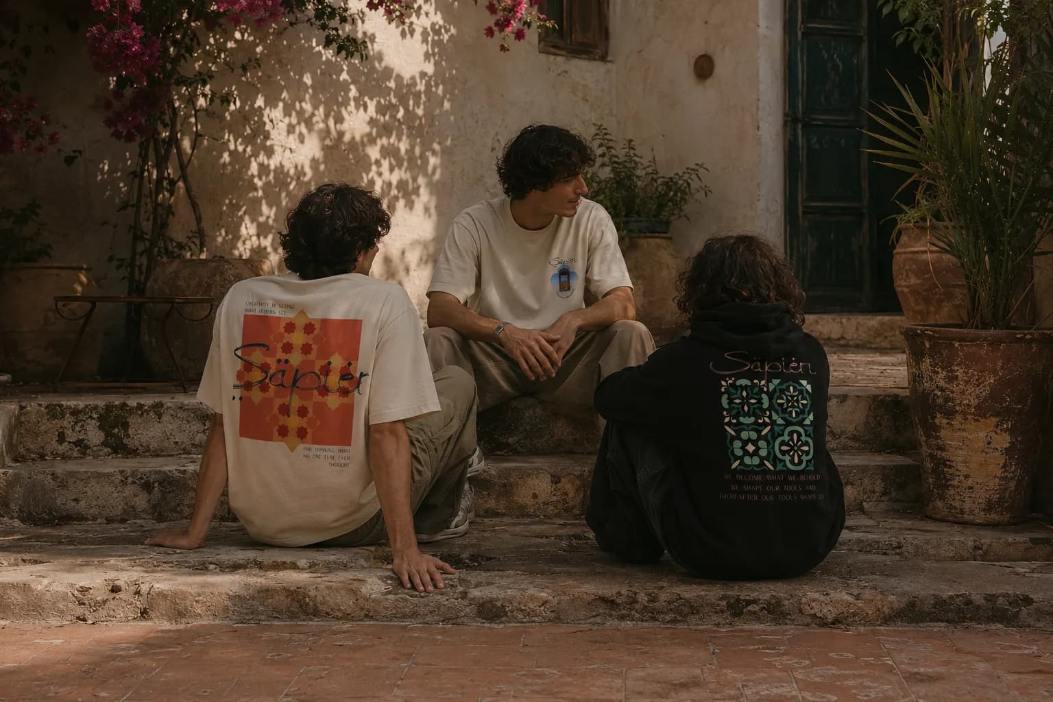 Three people sit on courtyard steps wearing the Einstein tee, the Churchill tee and the McLuhan hoodie