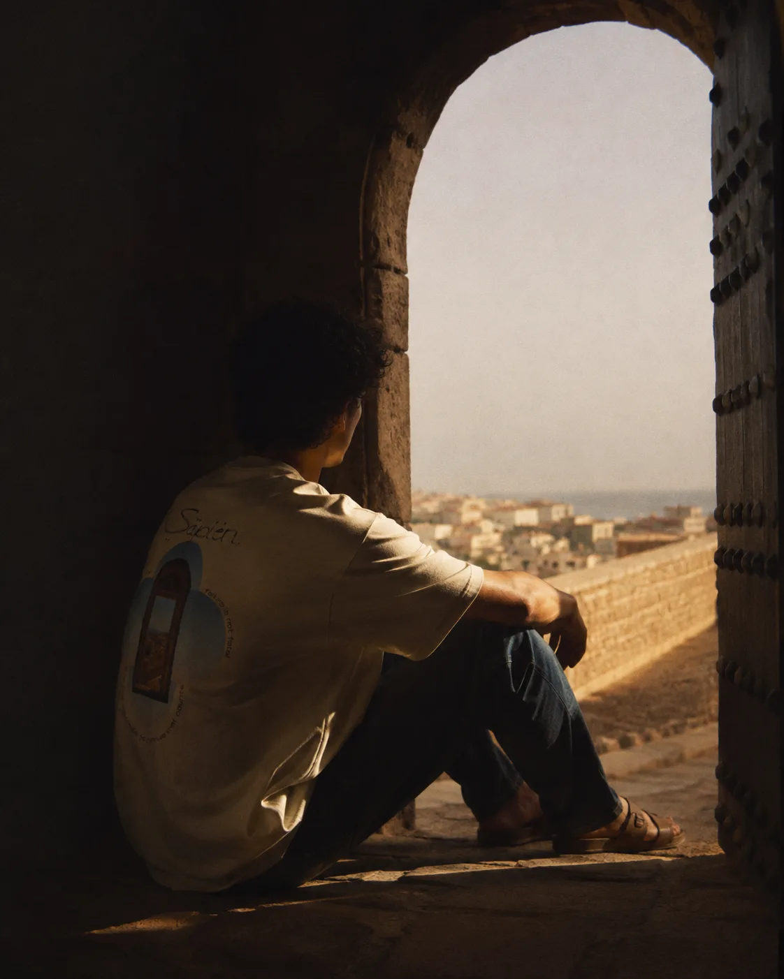 A man sits in a stone doorway above a Mediterranean hill town, wearing the Winston Churchill tee