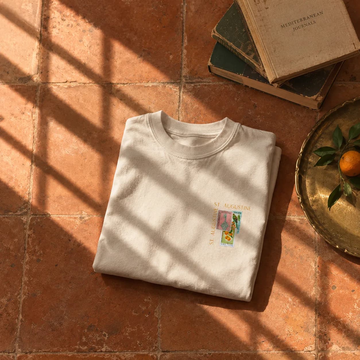 A folded bone tee resting on terracotta tile beside old books and a brass tray of oranges