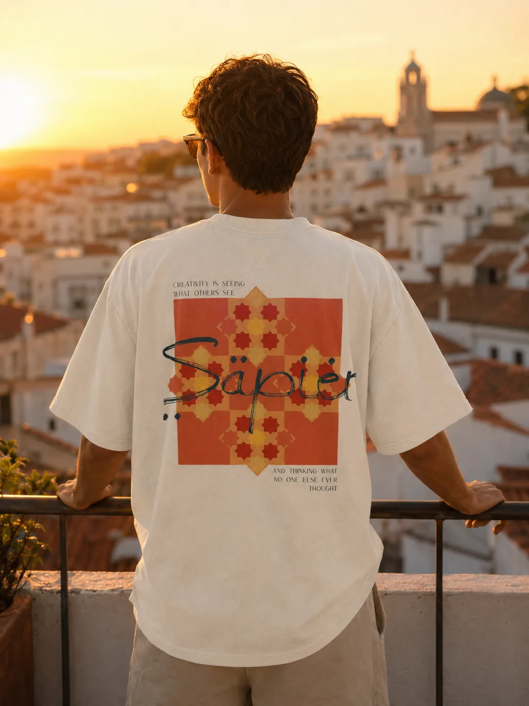 A man on a rooftop at sunset above a Mediterranean hill town, wearing the Einstein tee