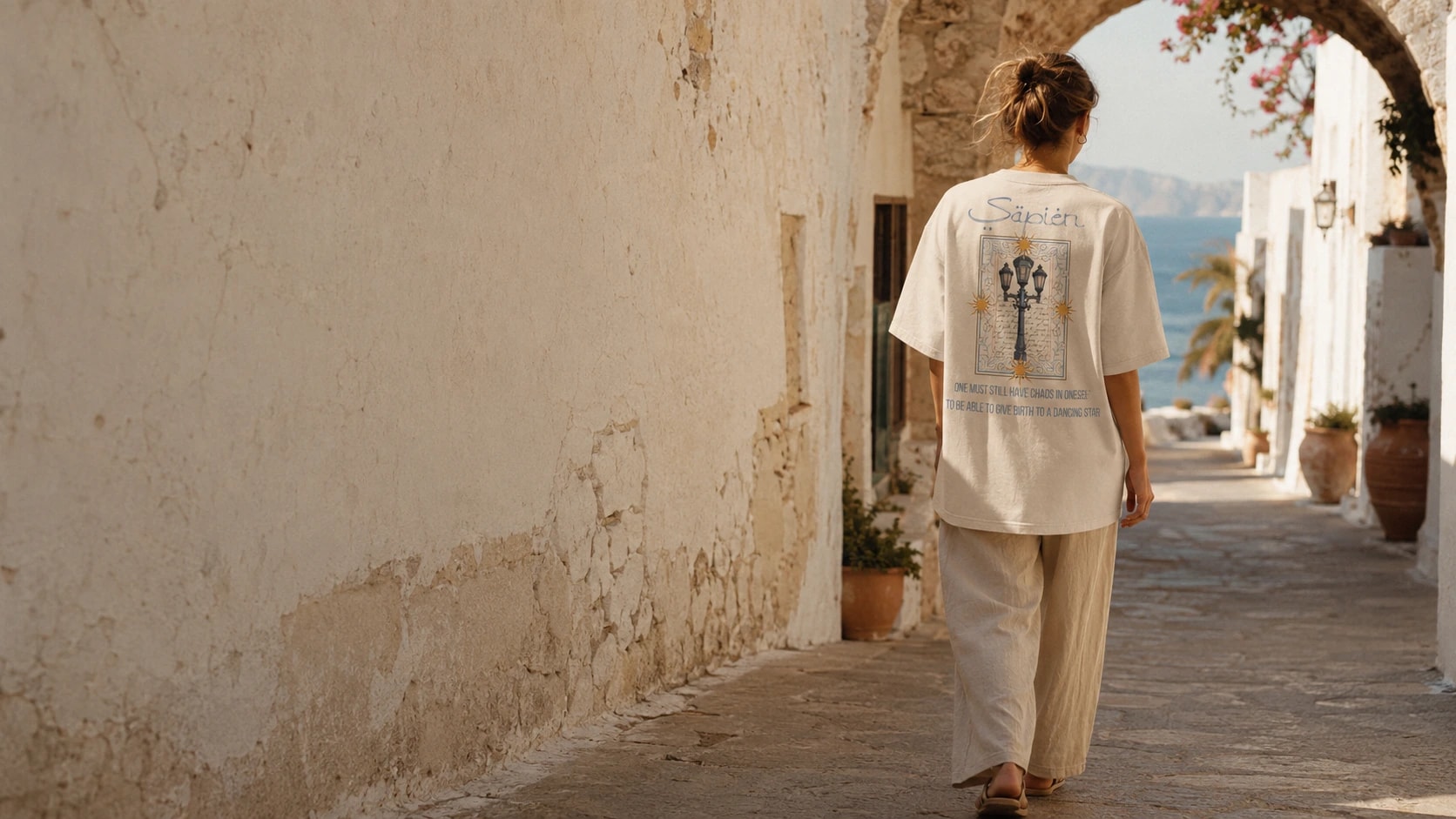 A woman walks away down a sunlit alley towards an archway, wearing a bone Sapien tee