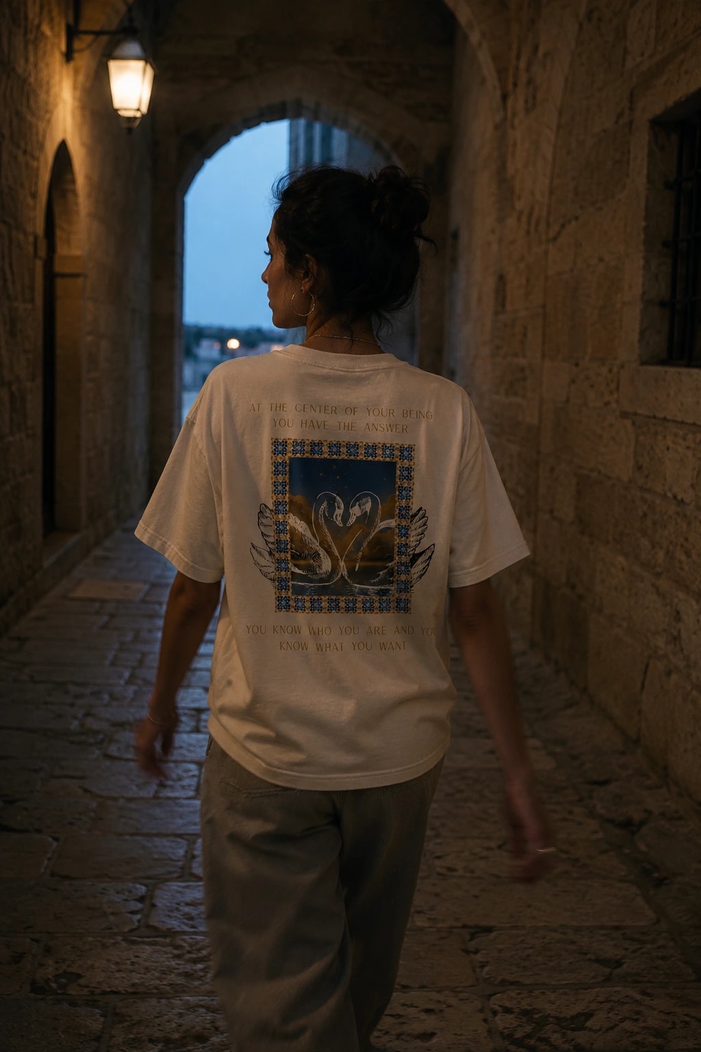 A woman walks through a lamplit stone archway at dusk wearing the Lao Tzu tee