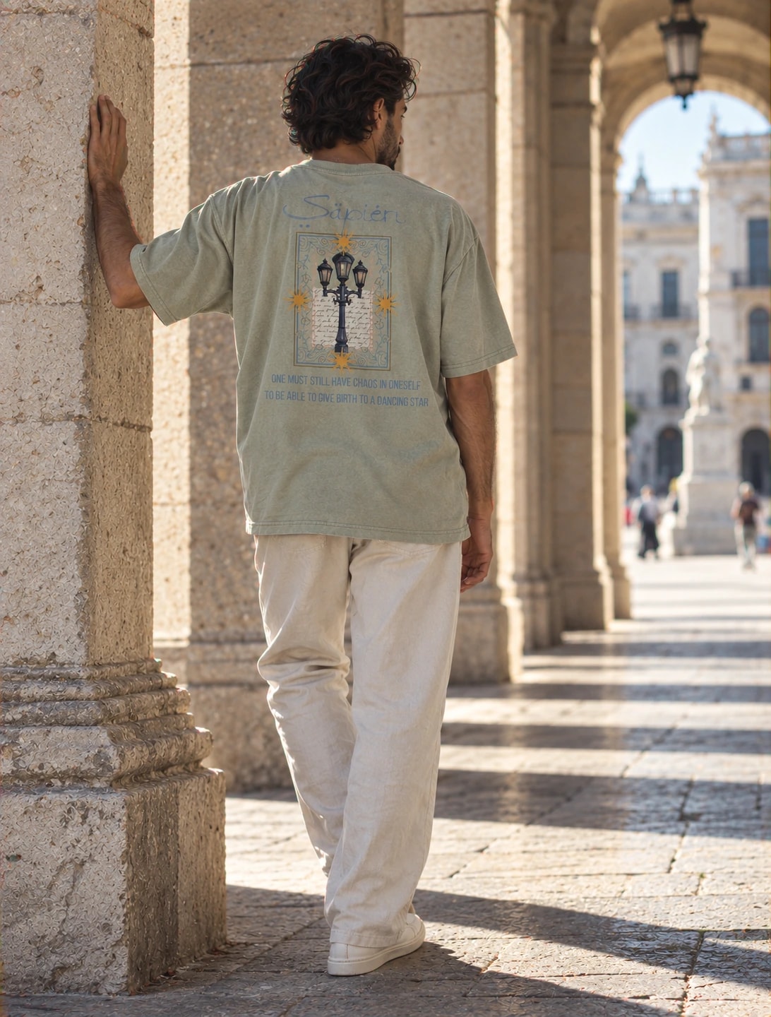 A man rests a hand on a stone column in a sunlit colonnade, wearing the Nietzsche tee, a plaza beyond the arch
